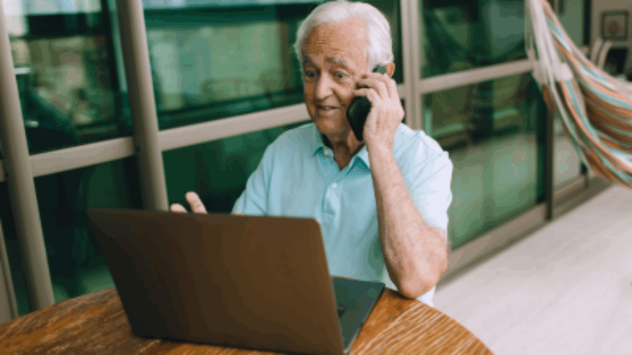 Senior siedzi przy komputerze i rozmawia przez telefon